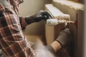 Home Service Blogs | Levine &Amp; Sons Plumbing, Heating, &Amp; Cooling A Person Wearing Gloves And A Plaid Shirt Adjusts Plumbing Fixtures On A Radiator, Demonstrating Expertise That Extends To Tasks Like Dehumidifier Repair.