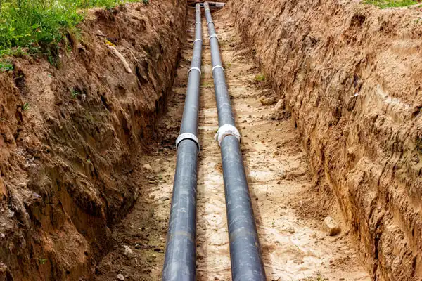Two long black pipes, part of a sewer line installation, are laid in a narrow, excavated trench and surrounded by soil.