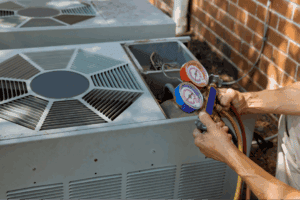 Home Service Blogs | Levine &Amp; Sons Plumbing, Heating, &Amp; Cooling A Person Uses A Manifold Gauge To Check The Pressure On An Outdoor Air Conditioning Unit Next To A Brick Wall, Showcasing Expert Heating Services In Southeastern Michigan.
