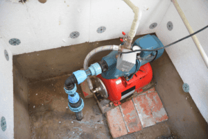 Home Service Blogs | Levine &Amp; Sons Plumbing, Heating, &Amp; Cooling A Water Pump System With Blue And Black Pipes, Installed In A Corner On A Brick And Concrete Base, Showcases Professional Sump Pump Services Detroit, Surrounded By White Insulated Walls For Added Protection.